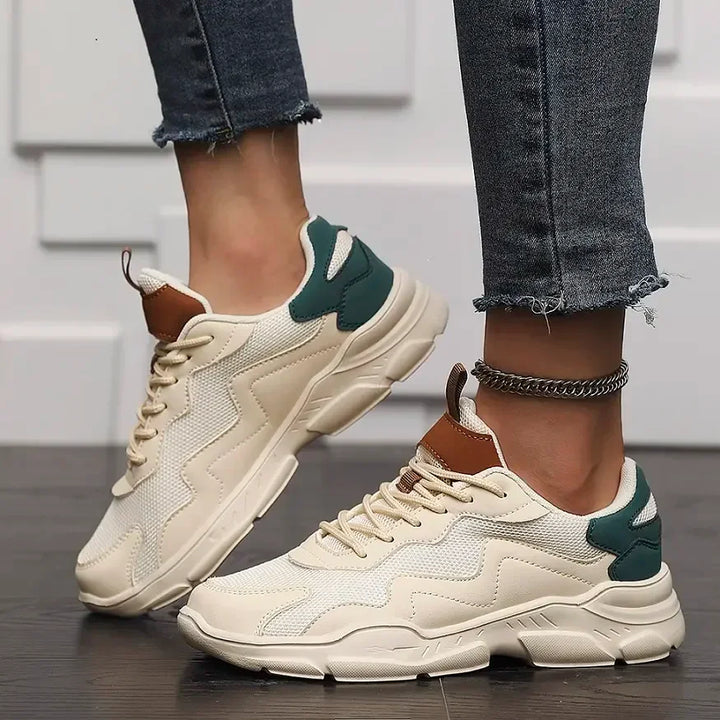 A person wearing beige chunky slip-on street shoes with green accents and brown details, featuring advanced shock absorption, paired with frayed-edge skinny jeans and a silver anklet, standing on a dark wooden floor against a white textured wall.