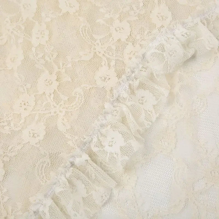 Close-up of cream-colored silky lace fabric with delicate patterns and a gathered ruffled edge running diagonally across the image, evoking an asymmetrical hemline.