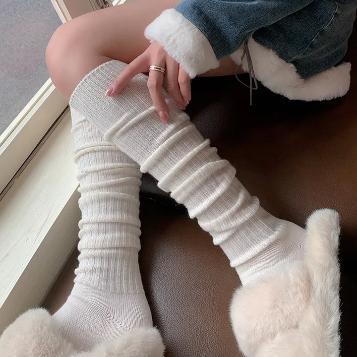 A person wearing cozy, breathable socks and fluffy slippers sits by a window on a brown seat, dressed in a denim jacket with white fur trim, gently touching one sock made from a soft Cashmere cotton blend.