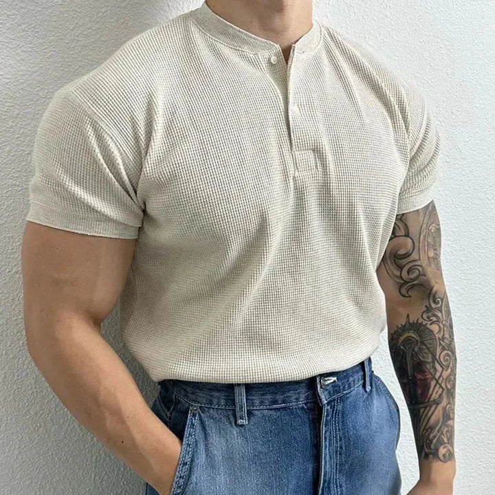 A person wearing a light beige, short-sleeve, ribbed texture shirt with a buttoned collar and blue jeans stands against a white wall. The form-fitting shirt highlights their muscular build and detailed tattoo on their right forearm.