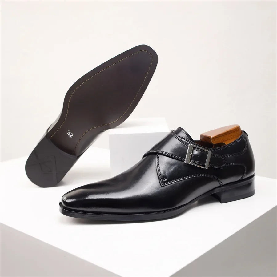 A pair of shiny black Italian leather monk strap shoes with a single buckle. One shoe is upright with a wooden shoe tree inside, while the other is sole up, showing size 42—all displayed on white geometric pedestals.