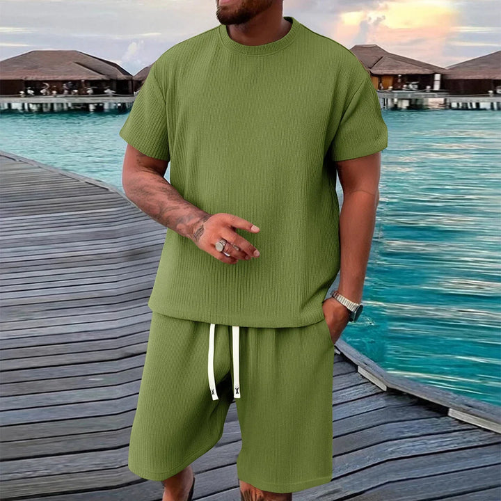 Man in olive green short sleeve shirt and shorts set on boardwalk by turquoise water, Rovelli style
