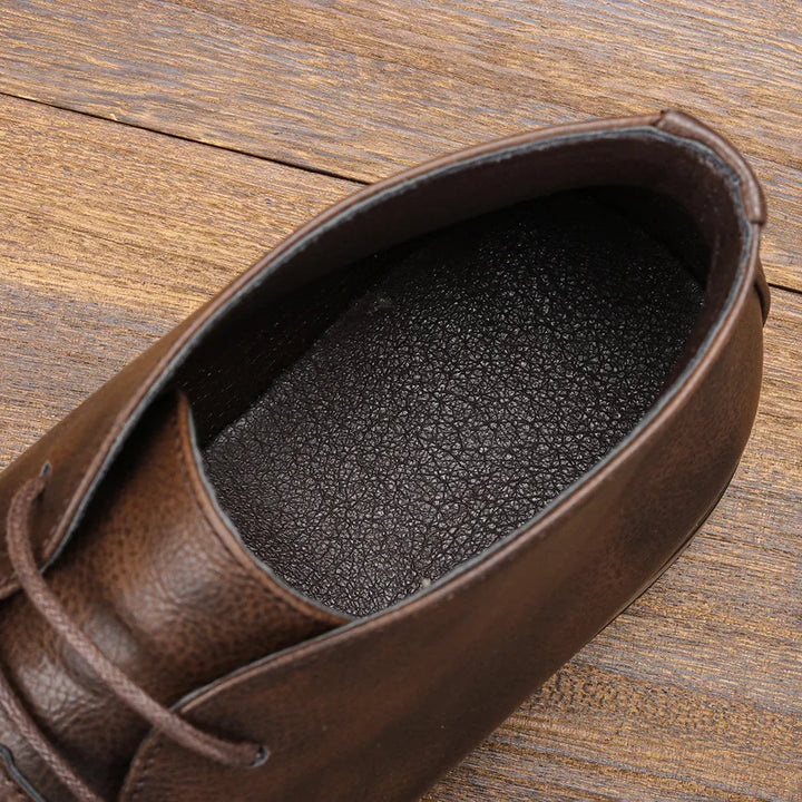 Close-up of the inside of a brown leather Western-inspired chukka, with visible laces, textured insole, and a wooden floor background.