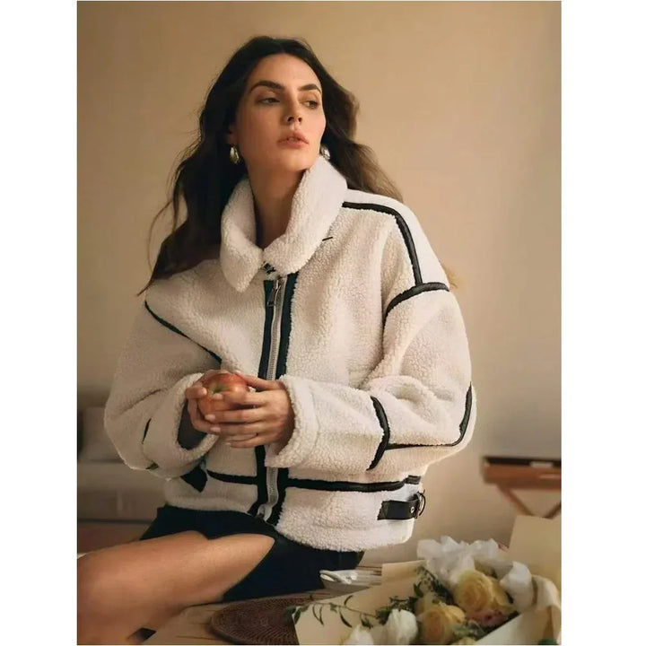 Woman in white shearling jacket with black trim sitting indoors, holding an apple, Rovelli fashion