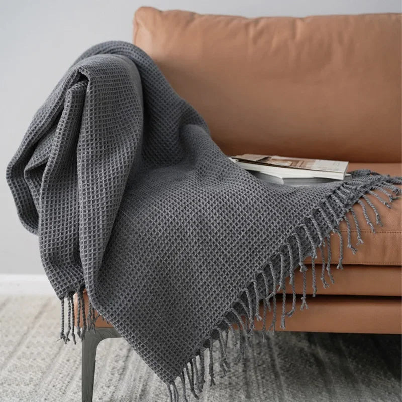 A gray knit waffle blanket with fringed edges is draped over a tan sofa, serving as a cozy decorative accent blanket. A book and magazine rest on the throw, creating an inviting scene against a light wall and patterned rug.