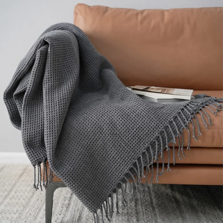 A gray knit waffle blanket with fringed edges is draped over a tan sofa, serving as a cozy decorative accent blanket. A book and magazine rest on the throw, creating an inviting scene against a light wall and patterned rug.