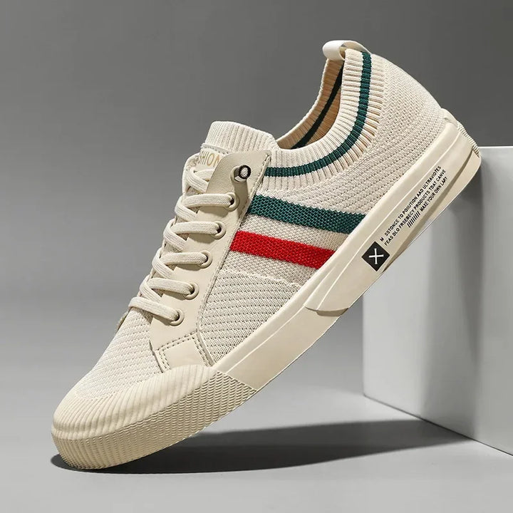 A beige knit sneaker with a breathable mesh upper, white laces, a ribbed sole, and green and red stripes on the side, leaning against a white surface on a gray background.