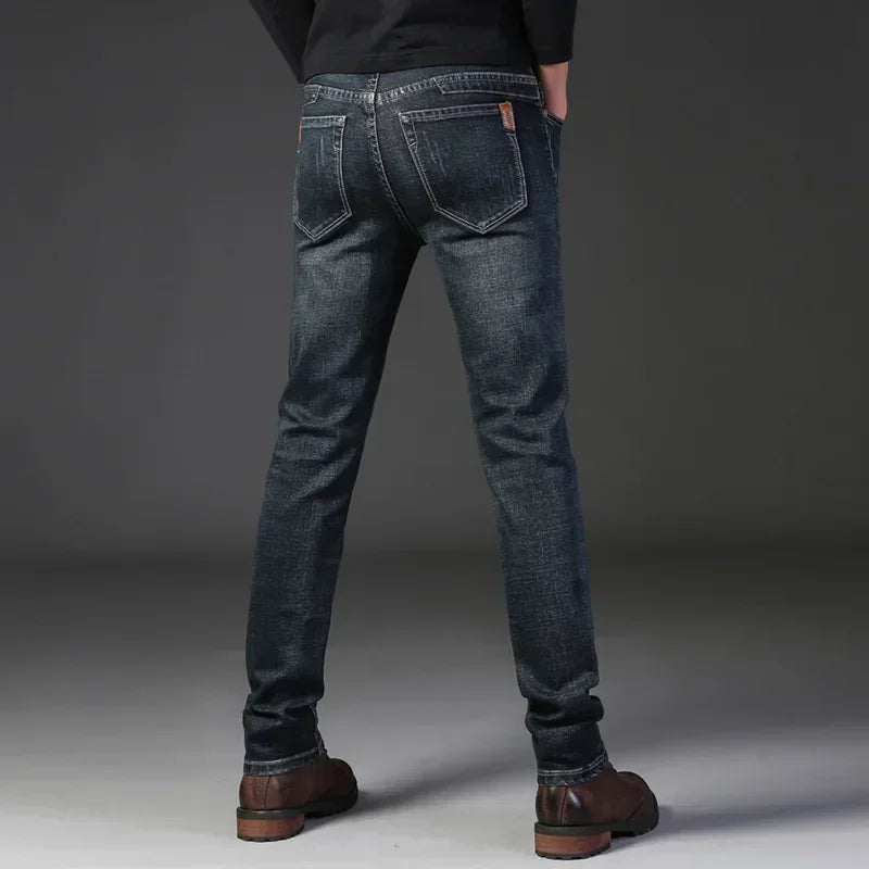 A person stands facing away, wearing slim-fit jeans made from durable fabric, brown shoes, and a black top. Their hands are in their pockets against a plain gray background.