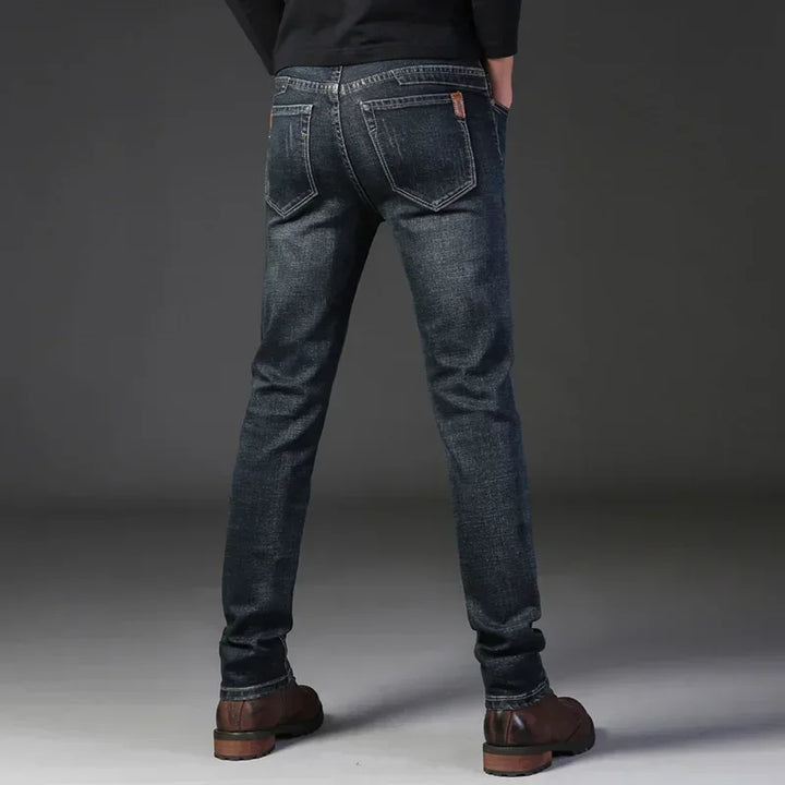 A person stands facing away, wearing slim-fit jeans made from durable fabric, brown shoes, and a black top. Their hands are in their pockets against a plain gray background.
