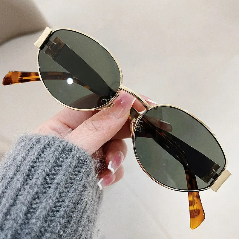 A hand wearing a gray knit sweater holds a pair of Italian style, oval-shaped sunglasses with gold frames, dark lenses, and tortoiseshell-patterned arms against a neutral background.