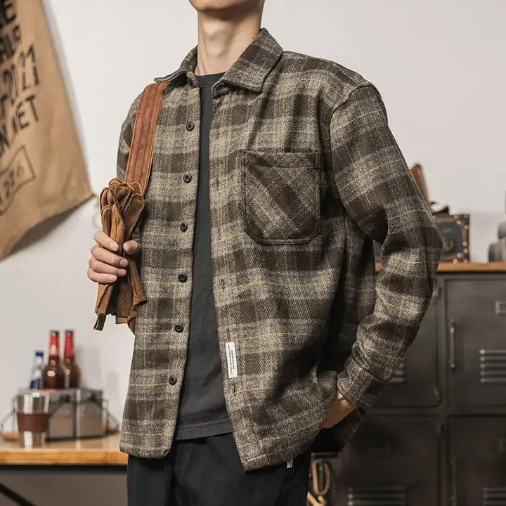 A person wearing a brown and gray plaid relaxed fit button-up shirt stands indoors with one hand in their pocket, holding a brown bag strap over their shoulder. The background shows bottles, lockers, and various items on a table.