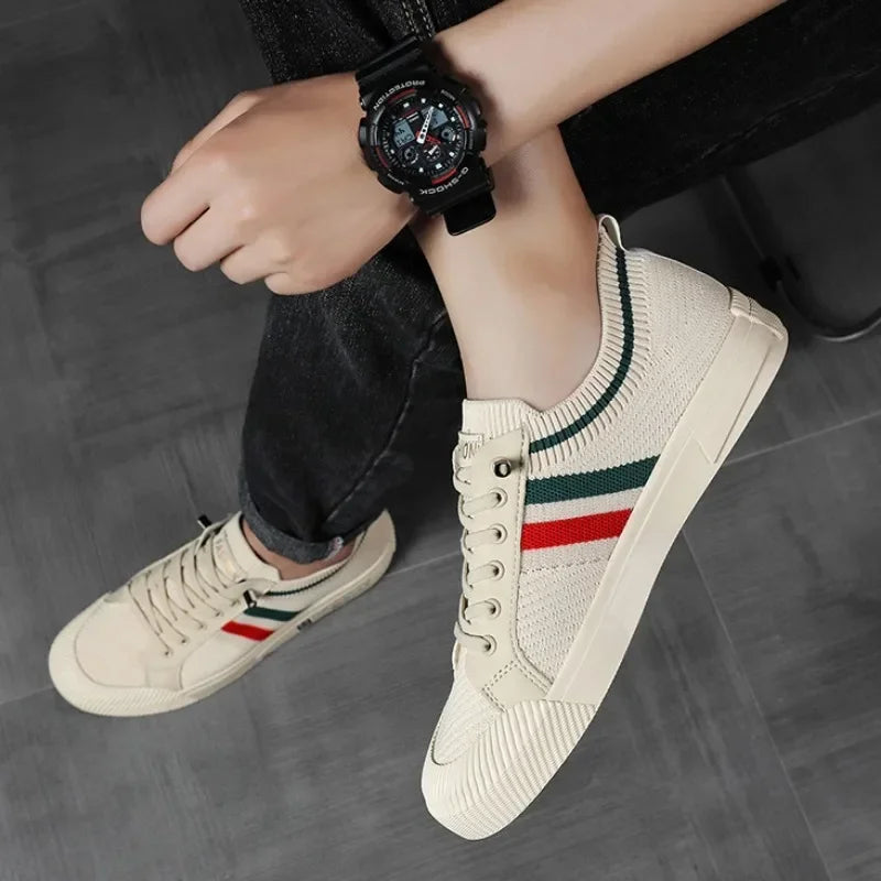 A person wearing cream-colored sneakers with a breathable mesh upper, green and red stripes, and a black wristwatch sits on a gray floor, with one leg crossed over the other.