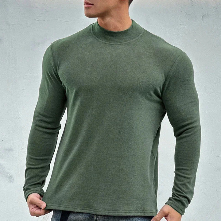 A person wearing a close-fitting, long-sleeve, olive green shirt with a mock neck collar and blue jeans stands against a plain light gray background, facing slightly to the side.