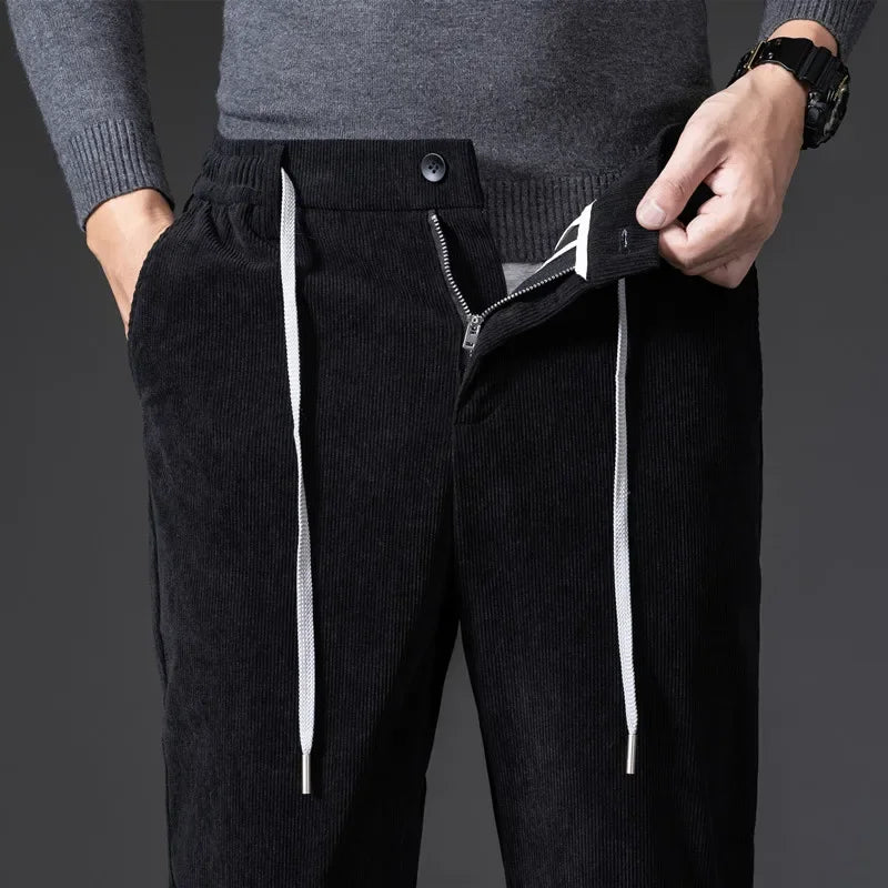 A person wearing men's smart casual corduroy pants with a zipper fly, a gray sweater, and a black watch is unbuttoning and unzipping the pants with one hand while the other hand is in their pocket.