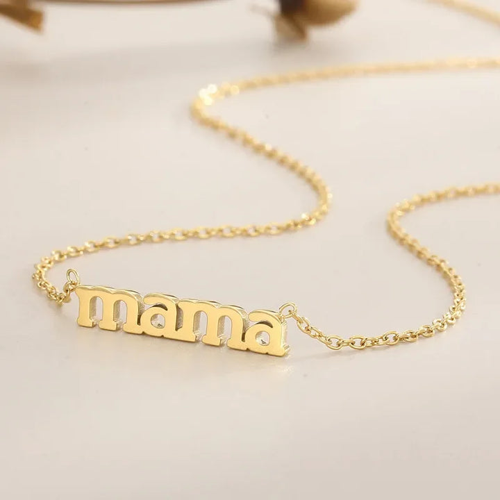 A close-up of a gold Mama Forever Necklace with a pendant spelling mama in lowercase letters, resting on a smooth, light-colored surface.