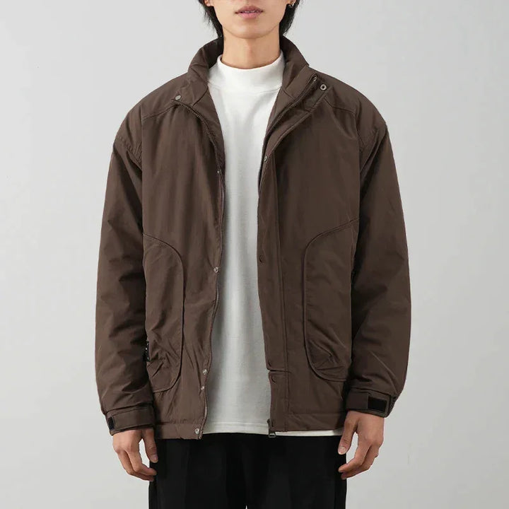 A person wearing a minimalist brown jacket with luxurious cotton lining over a white t-shirt and black pants stands against a plain light gray background. The jacket has a high collar and large front pockets.