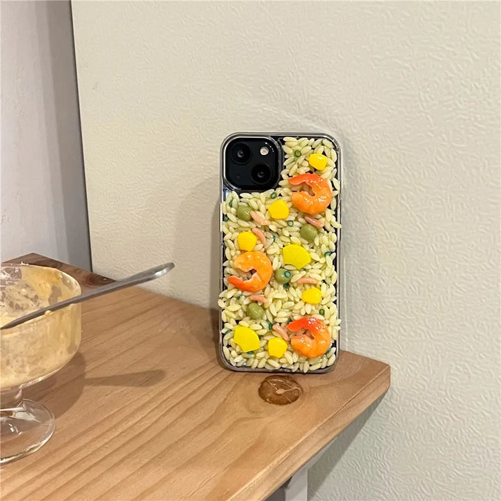 A phone with a shock-absorbent case designed to look like a shrimp salad, featuring realistic 3D detailing of shrimp, corn, and veggies. It’s propped against a pale wall beside a wooden surface with a glass dessert bowl and spoon.