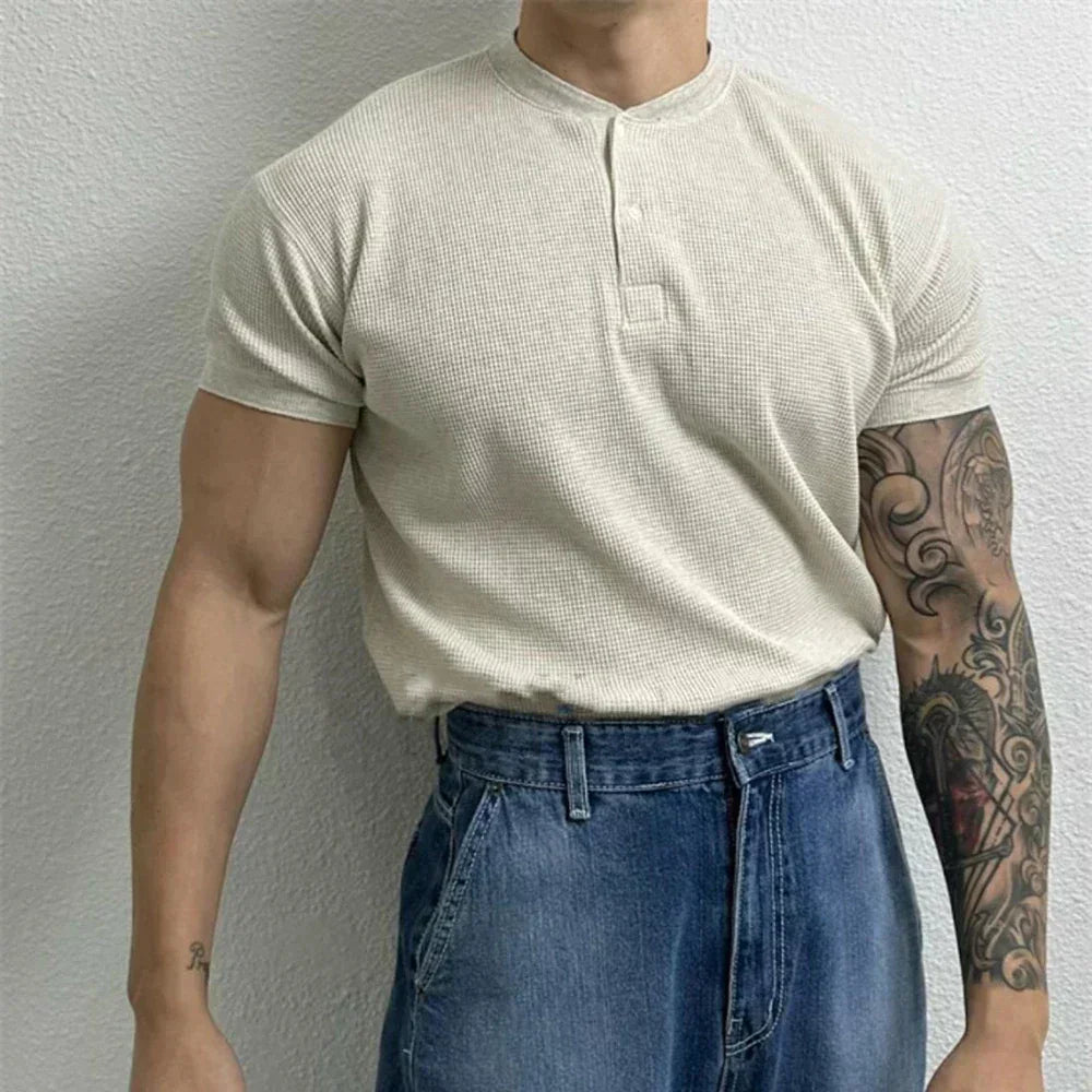 A person stands against a light-colored wall, wearing a ribbed Henley short-sleeve beige shirt tucked into high-waisted blue jeans. One arm has a colorful tattoo sleeve, while the other arm is bare.