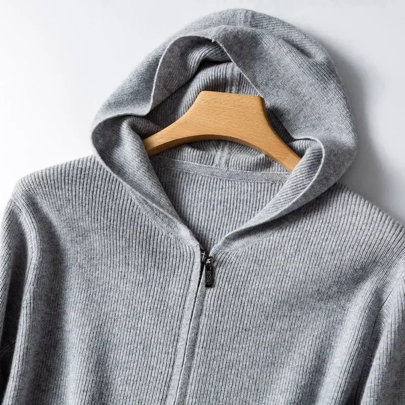 A close-up of a light gray, ribbed cashmere hoodie hanging on a wooden hanger against a white background. The hood is spread open, revealing the lightweight insulating texture of the fabric.