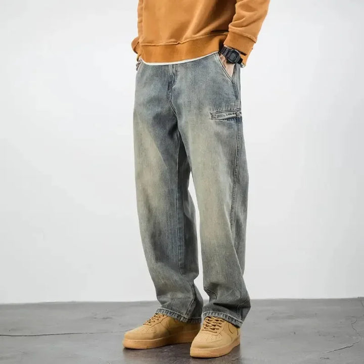 A person wearing a brown sweatshirt, classic fit stone washed jeans, and tan sneakers stands with hands in pockets against a plain, light background. Only the lower half of the body is visible.
