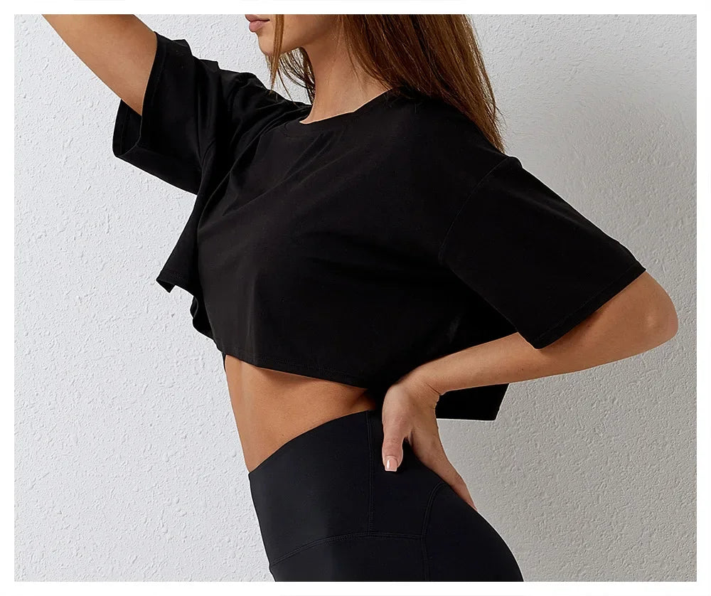 A woman poses in a cotton crop top and high-rise pants, standing sideways with one hand on her hip against a plain white background. Her long hair is draped over one shoulder.