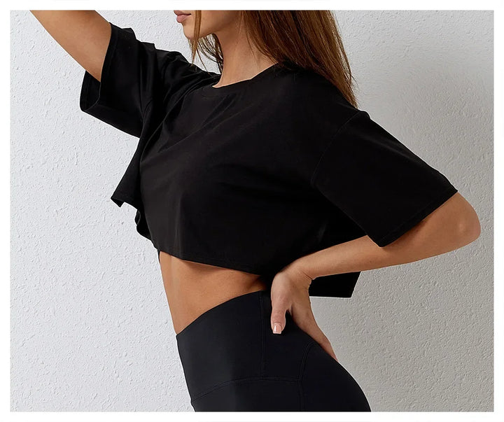 A woman poses in a cotton crop top and high-rise pants, standing sideways with one hand on her hip against a plain white background. Her long hair is draped over one shoulder.