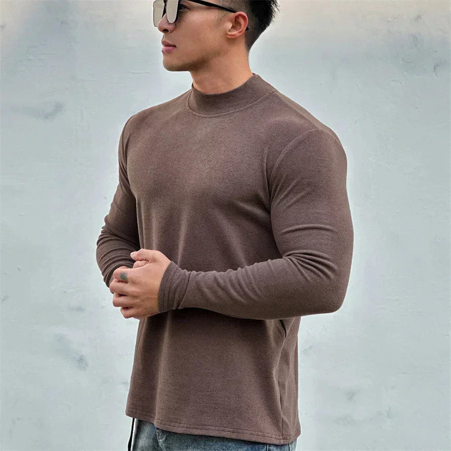 A man wearing sunglasses and a close-fitting brown long-sleeve shirt with a mock neck collar stands against a plain light gray background, holding his hands together in front of him.