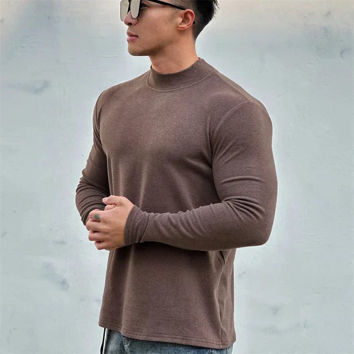 A man wearing sunglasses and a close-fitting brown long-sleeve shirt with a mock neck collar stands against a plain light gray background, holding his hands together in front of him.