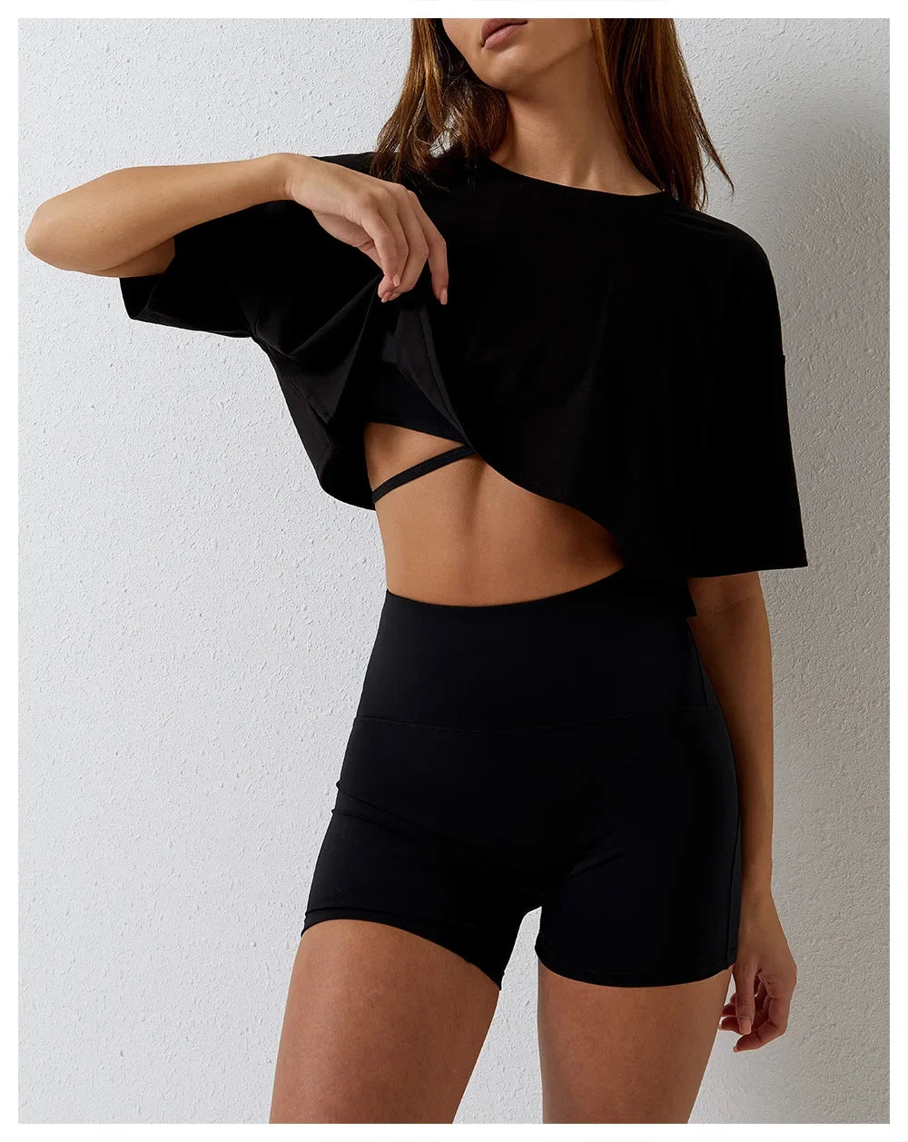 A woman wearing a relaxed fit black cotton crop top and high-waisted black bike shorts stands against a light-colored wall, lifting her shirt slightly to reveal a strappy detail underneath.