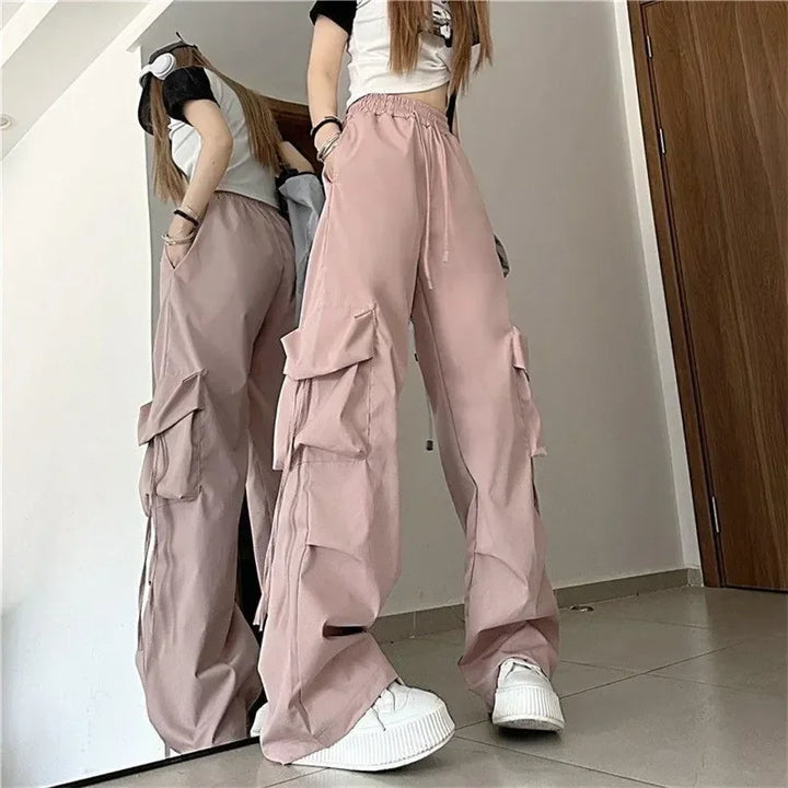 A person wearing loose, light pink cargo pants with a high-waist and multiple cargo pockets, a white t-shirt with black sleeves, and white sneakers stands indoors in front of a mirror, partially reflected.