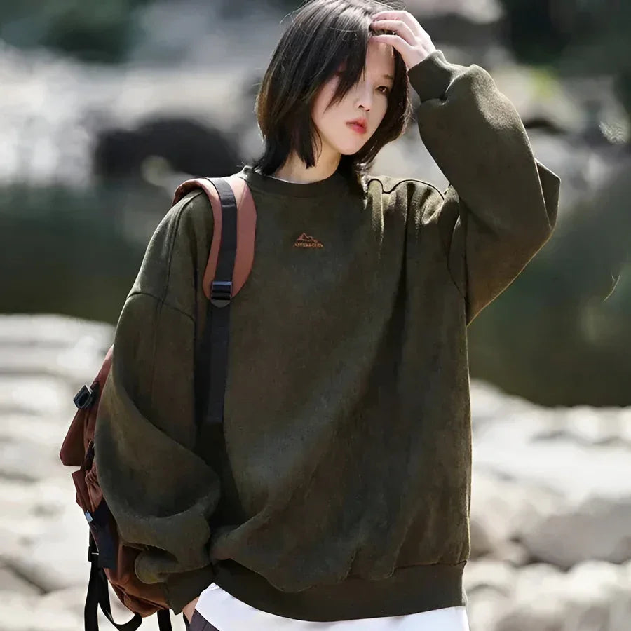 Woman outdoors in oversized green Rovelli sweatshirt with brown backpack, rocky background