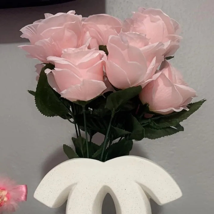 A bouquet of light pink artificial roses with green leaves in a Ceramic Vase, placed behind a white decorative object with a curved design, adds Nordic Home Decoration charm against a gray wall.