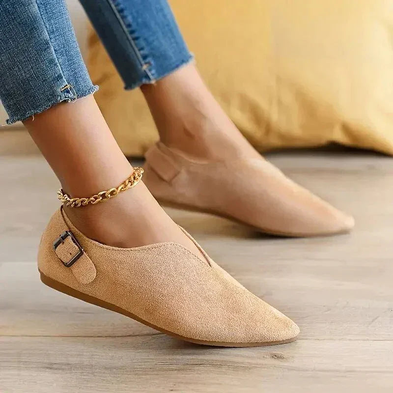 A person wearing light tan suede pointed toe shoes with a side buckle detail and cushioned insole, paired with blue frayed-hem jeans and a gold chain anklet, sitting indoors on a wooden floor near a yellow pillow.