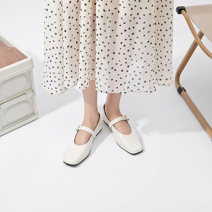 A person wearing a white, polka-dotted skirt and cream-colored sheepskin ladies shoes stands on a white floor near a beige foldable chair and a white storage box.