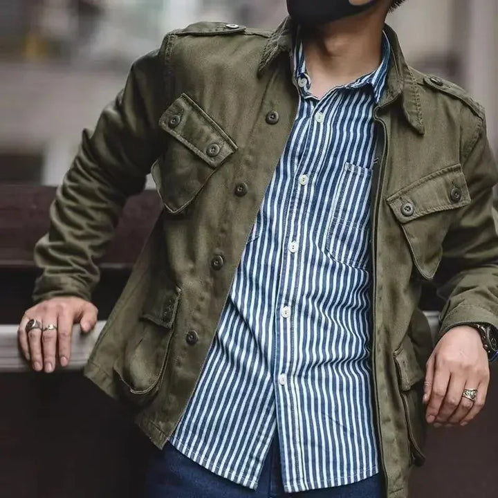 A person wearing a vintage military jacket over a blue and white vertically striped button-up shirt, dark jeans, and several rings, leans against a railing with their face partially out of frame.