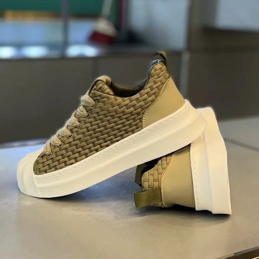 A pair of beige woven upper sneakers with thick white cushioned soles is displayed on a smooth surface; one shoe stands upright while the other is laid on its side beneath it.