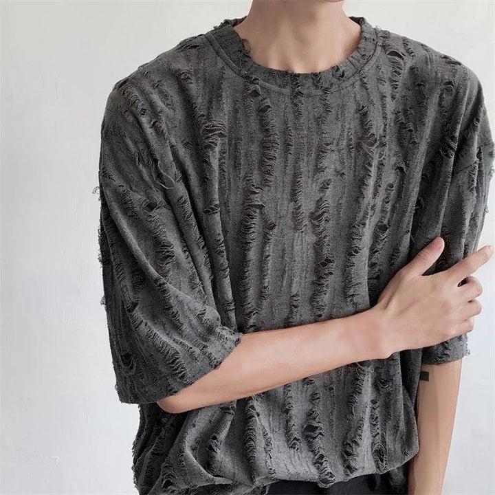 A person wearing a loose-fitting, dark gray, oversized tee made of distressed torn fabric with many rips and frayed edges stands against a plain light-colored background. Their left arm crosses their body, partially touching their right arm.