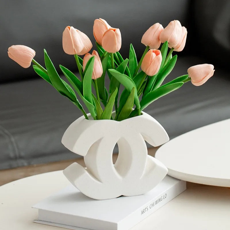 A white ceramic vase shaped like the Chanel logo holds pink tulips with green stems and leaves, placed on a closed book atop a table. Showcasing Irregular INS Style, it’s perfect for Nordic home decoration. A gray couch sits in the background.