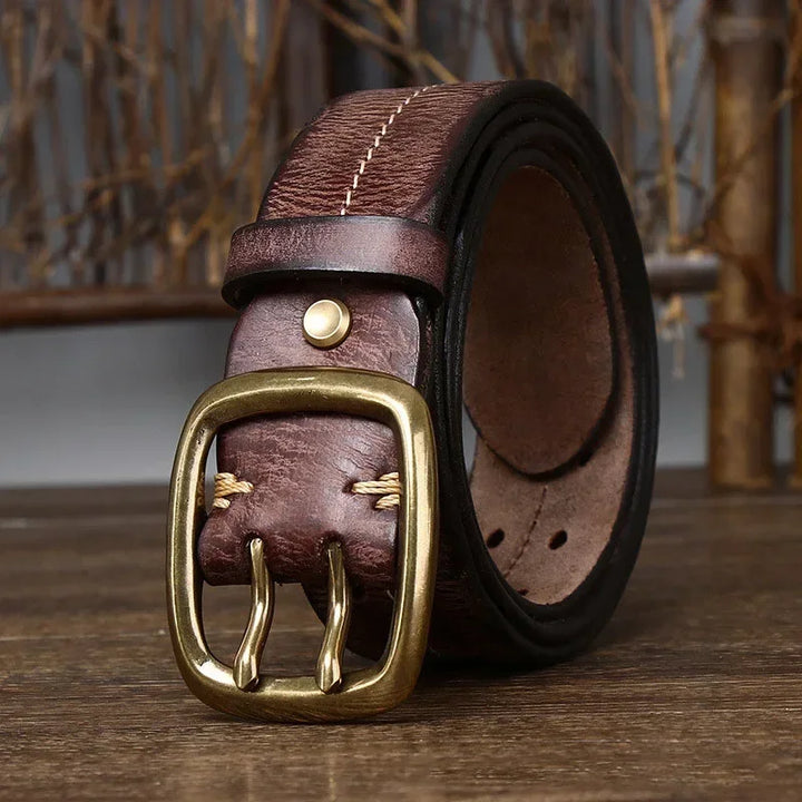 A rolled-up genuine leather belt with a brass double prong buckle, placed on a wooden surface with a blurred background of dried branches, showcases heritage craftsmanship.