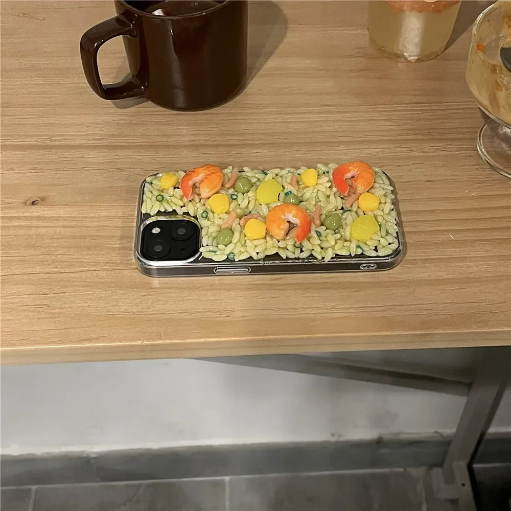 A smartphone with shock-absorbent, rugged phone protection features a case designed to look like a dish of rice, shrimp, corn, and peas. It sits on a wooden table next to a brown mug and a glass with a drink.