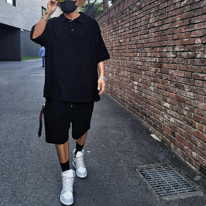 Man in oversized black shirt and shorts, white sneakers, walking on city street by brick wall