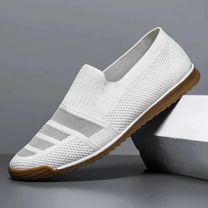 A white slip-on knit men's casual shoe with a textured upper, elastic band across the top, and a brown rubber sole. The breathable loafer is angled, resting on a gray block against a plain gray background.