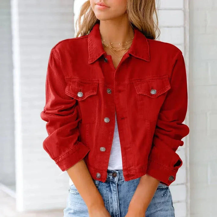 A woman wearing a bright red, cropped length denim jacket with silver buttons and chest pockets stands with her arms crossed over a white top and light blue jeans. Her wavy hair is loose as she poses in front of a white brick wall.