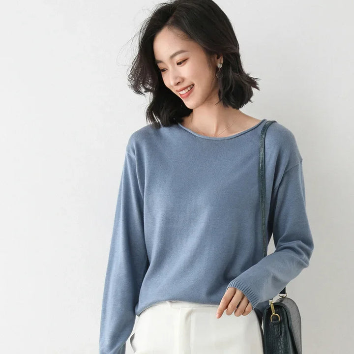 A woman with shoulder-length dark hair smiles while looking down. She is wearing a relaxed fit, light blue long-sleeve top, white pants, and carrying a dark shoulder bag. The background is plain and light-colored.