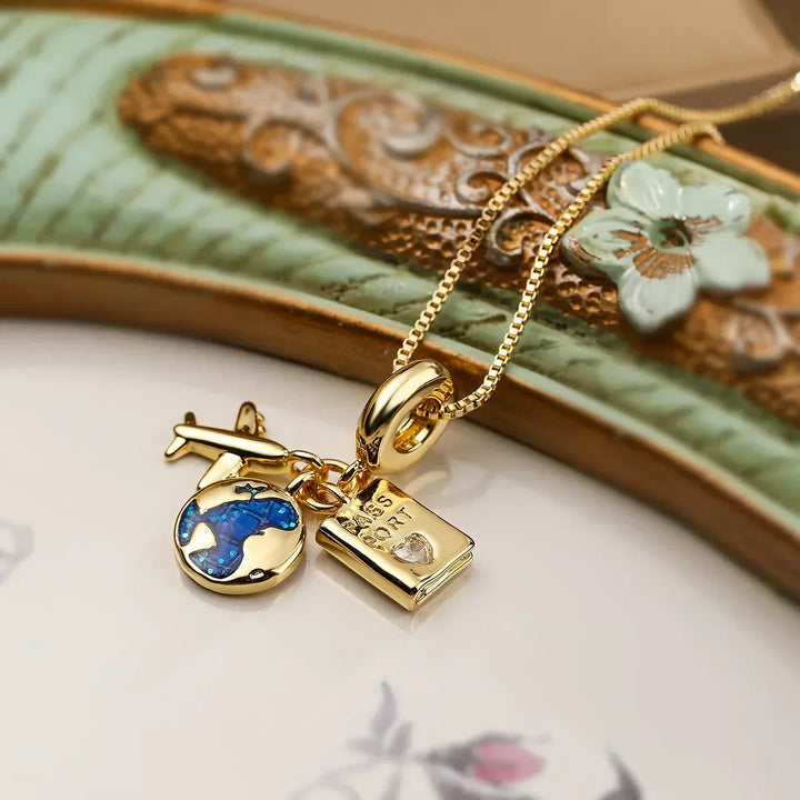 A gold adventure necklace with fine link craftsmanship, featuring three charms—a blue and green globe, gold airplane, and gemstone-accented passport—rests on a decorative ceramic surface adorned with floral details.
