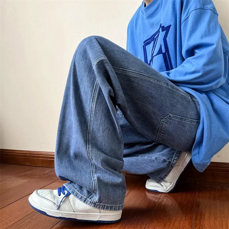 A person wearing relaxed fit blue jeans, a baggy blue sweatshirt with a graphic design, and white sneakers with blue laces is kneeling on a wooden floor against a plain wall.