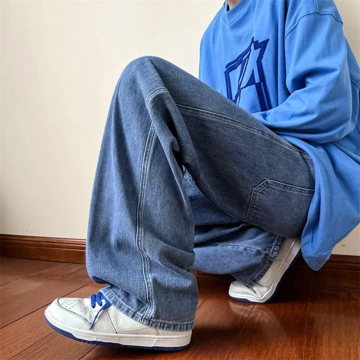 A person wearing relaxed fit blue jeans, a baggy blue sweatshirt with a graphic design, and white sneakers with blue laces is kneeling on a wooden floor against a plain wall.