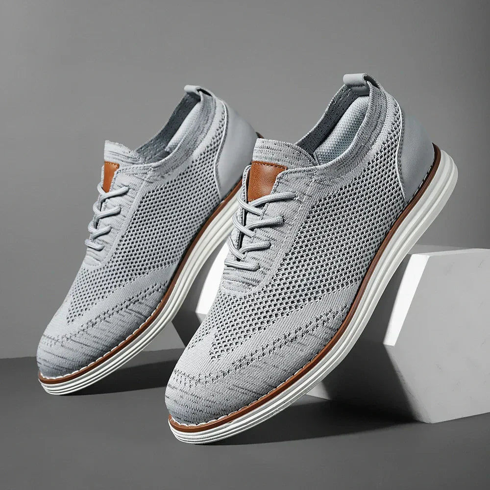 A pair of breathable knit sneakers in light gray with white soles and brown accents, displayed on a gray background with one shoe elevated on a white block.