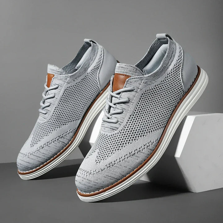 A pair of breathable knit sneakers in light gray with white soles and brown accents, displayed on a gray background with one shoe elevated on a white block.