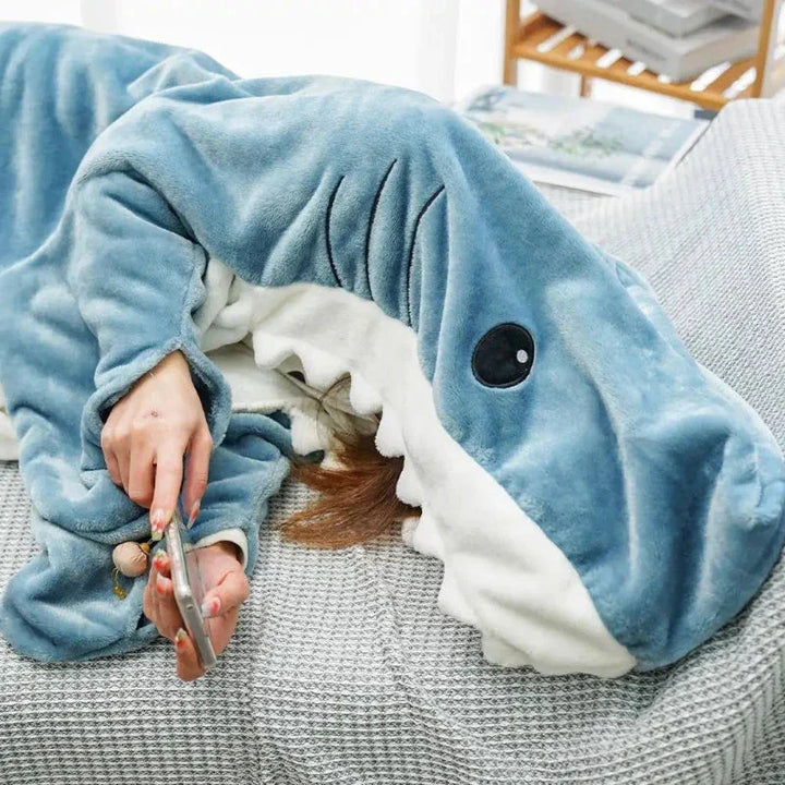 A person lying on a couch, mostly covered by a plush blue and white shark blanket with a large open mouth and black eyes, wearing cozy pajamas and holding a smartphone in one hand.